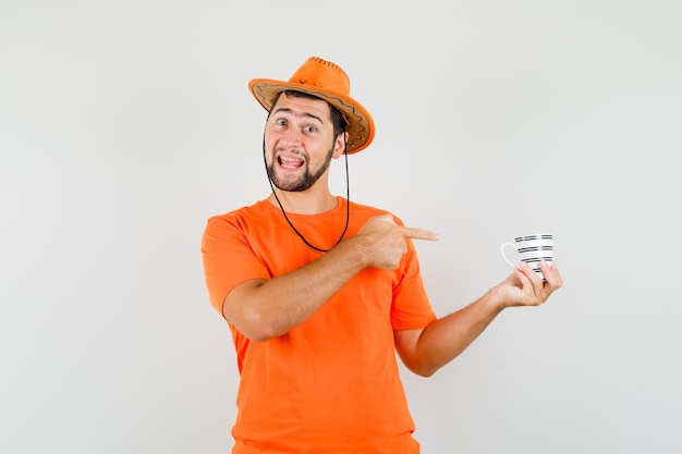 Giovane maschio che indica alla tazza di bevanda in maglietta arancione, cappello e guardando allegro, vista frontale.