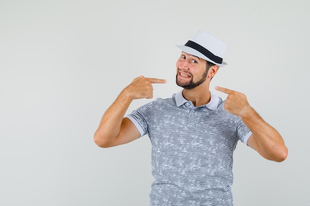 Giovane maschio che indica il suo sorriso in t-shirt, cappello e guardando allegro, vista frontale.