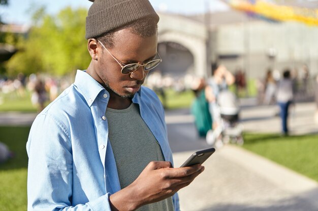 giovane maschio che indossa sfumature alla moda e camicia tenendo lo smartphone nelle mani