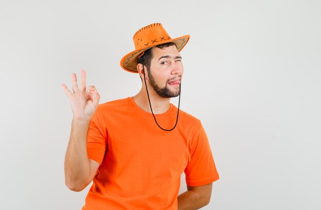 Giovane maschio che mostra gesto ok, sporge la lingua, strizza l'occhio in maglietta arancione, cappello, vista frontale.