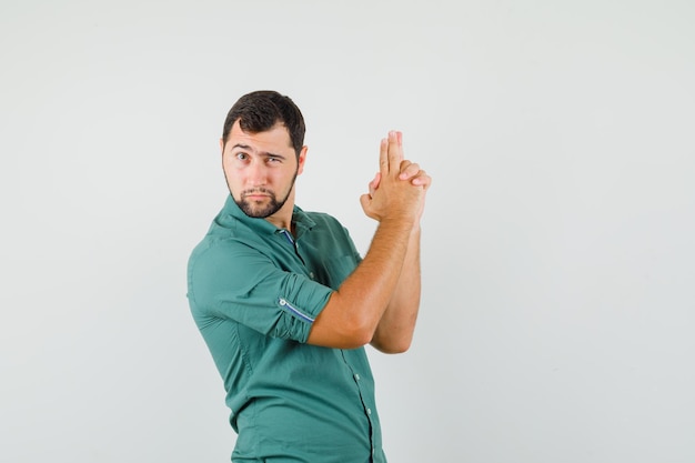 Giovane maschio che mostra il gesto della pistola di tiro in camicia verde e sembra coraggioso, vista frontale.