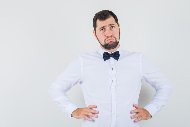 Giovane maschio che osserva in su con le mani sulla vita in camicia bianca e che sembra imbronciato. vista frontale.