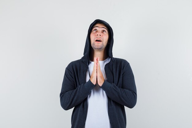 Giovane maschio che si tiene per mano nel gesto di preghiera in t-shirt, giacca e sembra speranzoso. vista frontale.