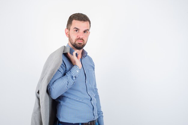 Giovane maschio che tiene giacca sulla spalla in camicia, jeans, giacca e sembra fiducioso, vista frontale.