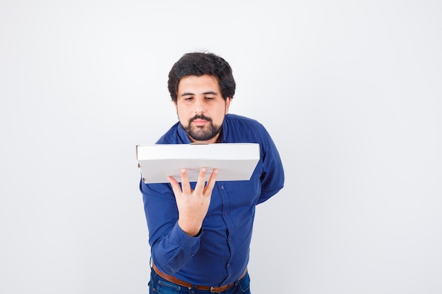 Giovane maschio che tiene la scatola della pizza in camicia, jeans e guardando fiducioso, vista frontale.