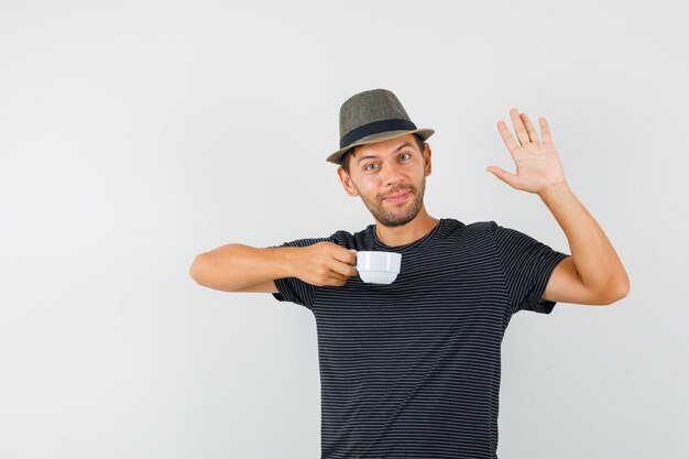 Giovane maschio che tiene tazza di bevanda agitando la mano in t-shirt hat e guardando allegro