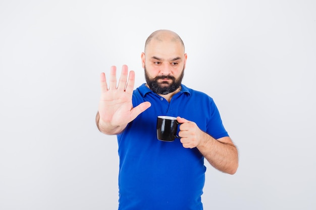 Giovane maschio che tiene tazza mentre mostra il gesto di arresto in camicia blu, vista frontale.