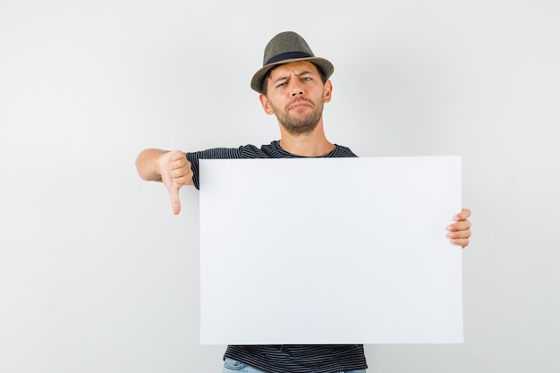 Giovane maschio che tiene tela di canapa in bianco che mostra il pollice giù in cappello dei jeans della maglietta