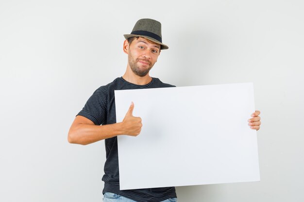 Giovane maschio che tiene tela in bianco che mostra il pollice in su in cappello dei jeans della maglietta e che sembra contento