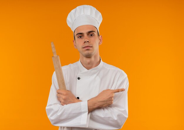 giovane maschio cool chef indossa uniforme incrocio mano che tiene il mattarello punta il dito a lato sul muro giallo isolato con spazio di copia