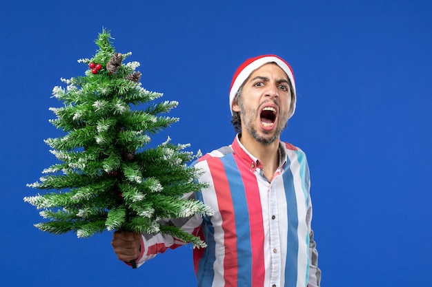 Giovane maschio di vista frontale arrabbiato con l'albero di nuovo anno sulla vacanza di emozione del nuovo anno della parete blu