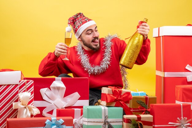 Giovane maschio di vista frontale che si siede intorno ai presente che celebra con champagne su fondo giallo