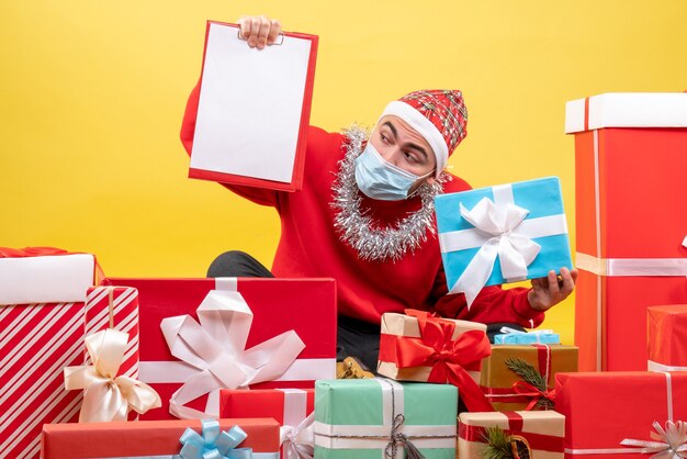 Giovane maschio di vista frontale che si siede intorno ai regali di Natale con la nota su fondo giallo