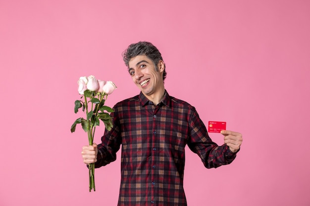 Giovane maschio di vista frontale che tiene belle rose rosa e carta di credito sulla parete rosa