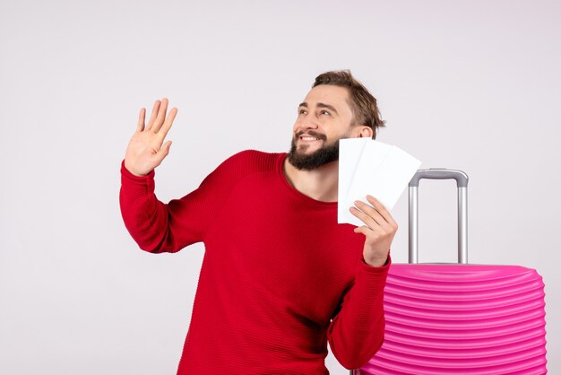 Giovane maschio di vista frontale con il sacchetto rosa e che tiene i biglietti aerei sul turista della foto di emozione di vacanza di volo di viaggio della parete bianca