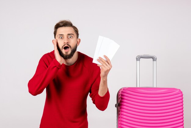 Giovane maschio di vista frontale con la borsa rosa e che tiene i biglietti sulla foto di vacanza turistica di viaggio di colore di volo di viaggio della parete bianca