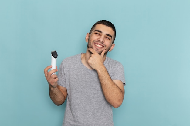 Giovane maschio di vista frontale in maglietta grigia che tiene il rasoio elettrico che sorride sulla schiuma dei capelli della barba da barba maschio blu ghiaccio