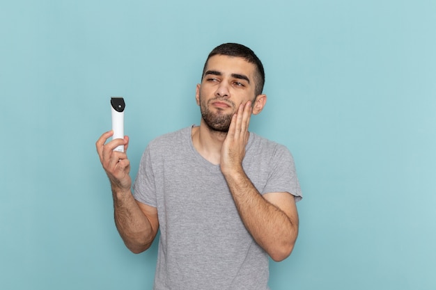 Giovane maschio di vista frontale in maglietta grigia che tiene il rasoio elettrico sulla schiuma dei capelli della barba da barba maschio blu ghiaccio