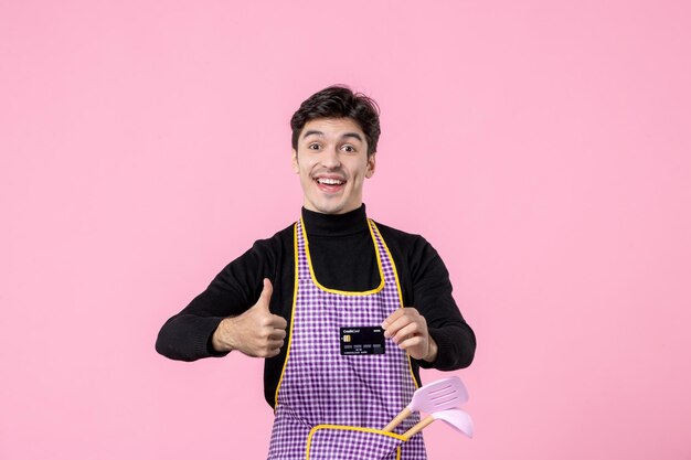 Giovane maschio di vista frontale in mantello che tiene carta di credito nera su sfondo rosa professione di lavoro che cucina il capo uniforme cucina colore denaro orizzontale