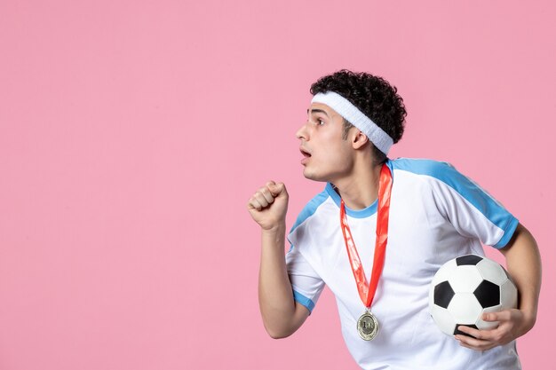 Giovane maschio di vista frontale in vestiti di sport con il pallone da calcio sulla parete rosa
