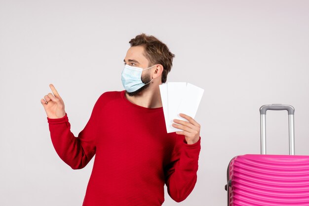 Giovane maschio di vista frontale nella maschera sterile che tiene i biglietti sul colore di viaggio dei voli del virus dell'emozione di vacanza di covid-aereo della parete bianca