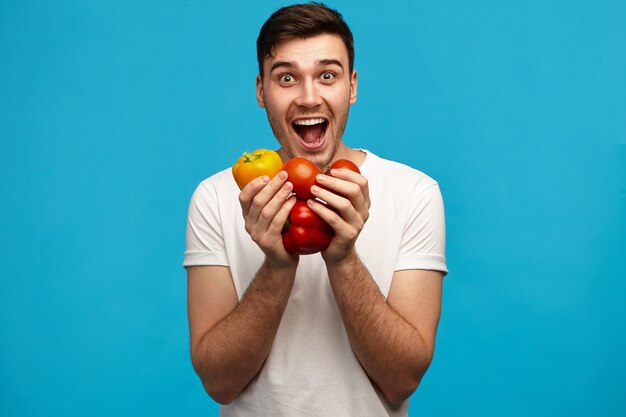 Giovane maschio emotivo divertente in camicia bianca che tiene peperoni e pomodori in entrambe le mani, con sguardo eccitato, aprendo ampiamente la bocca, felicissimo con verdure biologiche fresche dal suo giardino