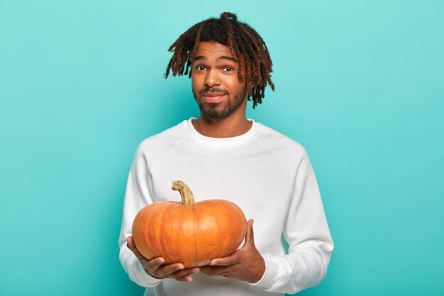 Giovane maschio giovane con i dreadlocks, poca barba, indossa un maglione bianco casual, tiene la zucca arancione, si prepara per la celebrazione delle vacanze