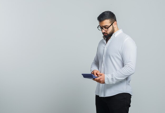 Giovane maschio in camicia bianca, pantaloni utilizzando la calcolatrice e guardando occupato