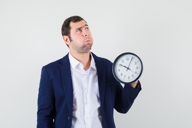 Giovane maschio in camicia e giacca tenendo l'orologio da parete e guardando pensieroso