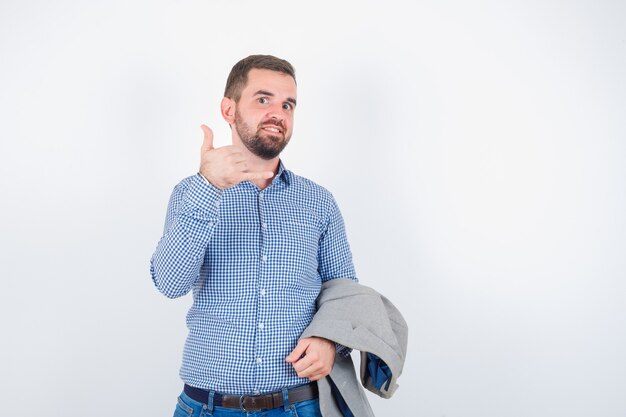 Giovane maschio in camicia, jeans, giacca da abito che tiene la giacca mentre mostra il gesto del telefono e sembra felice, vista frontale.