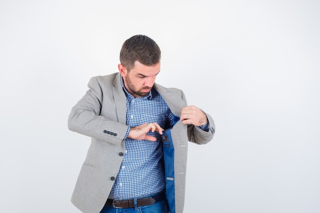 Giovane maschio in camicia, jeans, giacca da abito guardando la tasca della giacca e guardando serio, vista frontale.