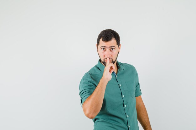 Giovane maschio in camicia verde che mostra gesto di silenzio e sembra concentrato, vista frontale.