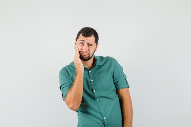 Giovane maschio in camicia verde che soffre di mal di denti e sembra doloroso, vista frontale.