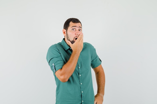 Giovane maschio in camicia verde che tiene la mano sulla bocca e sembra sorpreso, vista frontale.