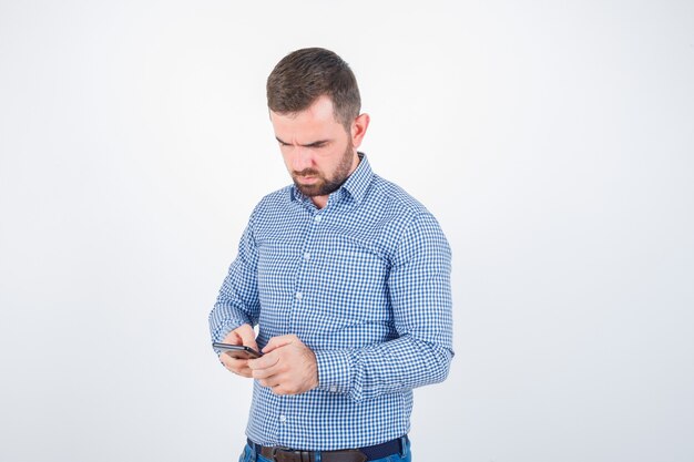 Giovane maschio in chat sul telefono cellulare in camicia, jeans e guardando pensieroso, vista frontale.