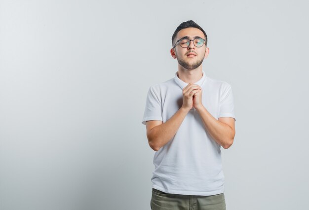 Giovane maschio in maglietta bianca, pantaloni stringendo le mani nel gesto di preghiera e guardando speranzoso