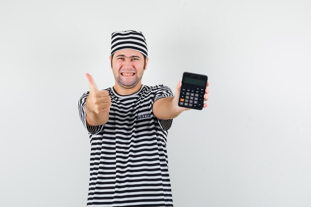 Giovane maschio in maglietta, cappello che mostra la calcolatrice con il pollice in su e che sembra allegra, vista frontale.
