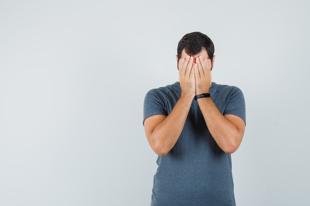 Giovane maschio in maglietta grigia mantenendo le mani sul viso e guardando depresso