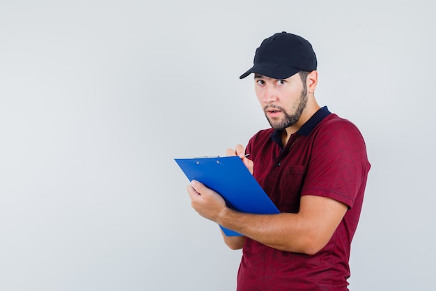Giovane maschio in maglietta rossa, berretto nero che guarda l'obbiettivo mentre scrive qualcosa sul suo taccuino e guarda attento, vista frontale.