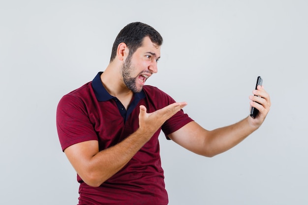 Giovane maschio in maglietta rossa che grida mentre guarda il telefono e sembra arrabbiato, vista frontale.