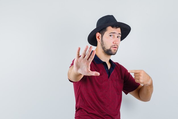 Giovane maschio in t-shirt, cappello che mostra il gesto di arresto mentre indica se stesso e sembra ansioso, vista frontale.