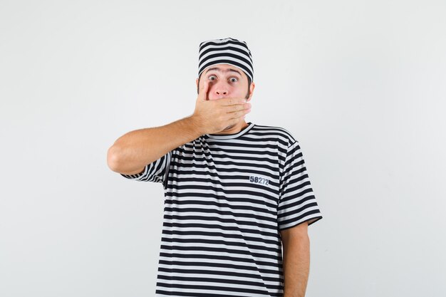Giovane maschio in t-shirt, cappello tenendo la mano sulla bocca e guardando scioccato, vista frontale.