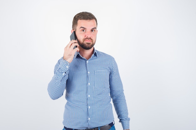 Giovane maschio parlando al cellulare in camicia, jeans e guardando fiducioso, vista frontale.