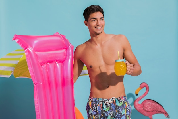 Giovane maschio sorridente che posa con il materasso di nuoto e della bevanda in studio