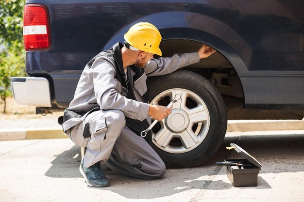 Giovane meccanico in abiti da lavoro e elmetto protettivo giallo utilizzando la chiave per cambiare la ruota dell'auto all'aperto