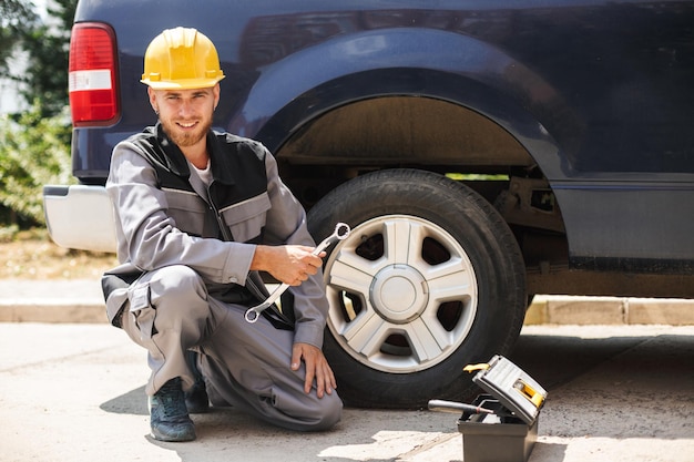 Giovane meccanico sorridente in abiti da lavoro e elmetto protettivo giallo che guarda felicemente nella macchina fotografica tenendo la chiave per cambiare la ruota dell'auto all'aperto