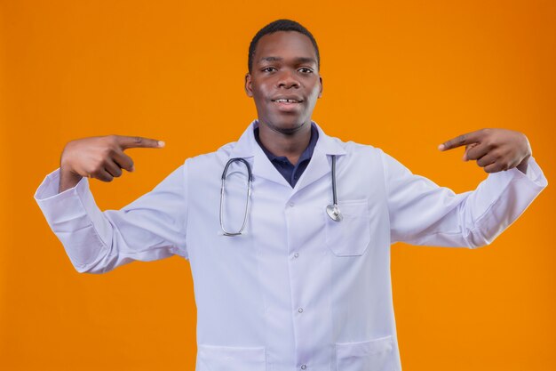 Giovane medico afroamericano che indossa camice bianco con lo stetoscopio che punta a se stesso con le dita indice sorridendo fiducioso