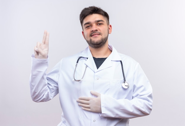 Giovane medico bello che indossa guanti medici bianchi dell'abito medico bianco e stetoscopio che tiene lo stomaco che mostra il segno di pace che sta sopra la parete bianca