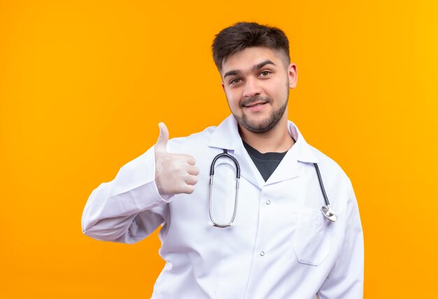 Giovane medico bello che indossa guanti medici bianchi e stetoscopio bianchi dell'abito medico che fanno i pollici felici in su che si leva in piedi sopra la parete arancione
