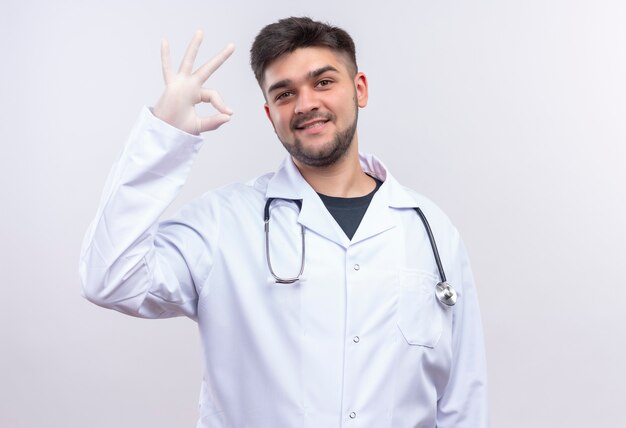 Giovane medico bello che indossa guanti medici bianchi e stetoscopio bianchi dell'abito medico che sorridono felicemente mostrando il segno giusto con il braccio che sta sopra la parete bianca